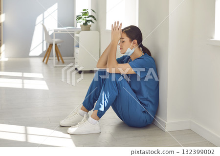 Tired stressed woman doctor wearing blue face mask and uniform sitting on hospital floor. 132399002