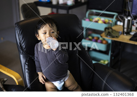 Toddler Drinking Milk Bottle On Office Chair, playful home office moment perfect for parenting ads, family lifestyle campaigns, and baby product promotions. 132401368