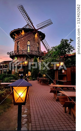 A stone building shaped like a windmill stands illuminated against a colorful twilight sky. It is not doing anything. The foreground shows an empty outdoor patio with wooden tables, benches, and warm, A stone building shaped like a windmill stands illuminated against a colorful twilight sky. It is not doing anything. The foreground shows an empty outdoor patio with wooden tables, benches, and warm, 132402062