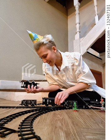 A happy woman in a party hat is sitting on the floor, playing with a black toy model train set. 132407101