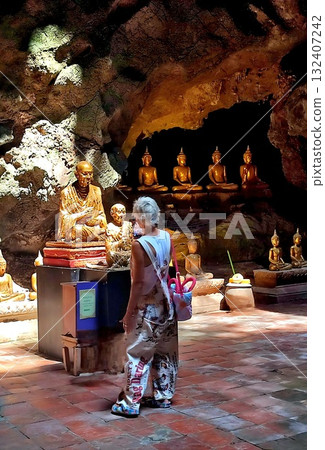 Tourist woman in patterned overalls stands inside a dimly lit Thai cave temple, looking at golden Buddha statues. 132407242