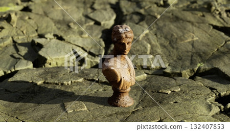 A small antique bust sculpture stands alone amidst cracked earth, revealing a stark contrast of detail against a rough surface. The bright daylight highlights its features and setting. 132407853
