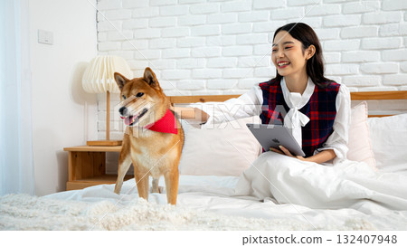 Asian woman smiling and petting her Shiba Inu dog while using a tablet on bed in cozy bedroom. Concept of pet companionship, digital lifestyle, relaxation, happiness, and work-from-home Asian woman smiling and petting her Shiba Inu dog while using a tablet on bed in cozy bedroom. Concept of pet companionship, digital lifestyle, relaxation, happiness, and work-from-home 132407948