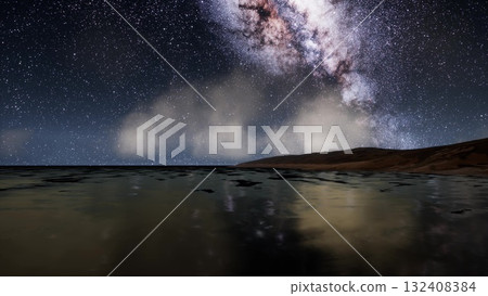 Under a canopy of stars, the tranquil sea reflects the wonders of the galaxy. Waves softly lap against the shore as the Milky Way illuminates the night sky, creating a breathtaking view. 132408384