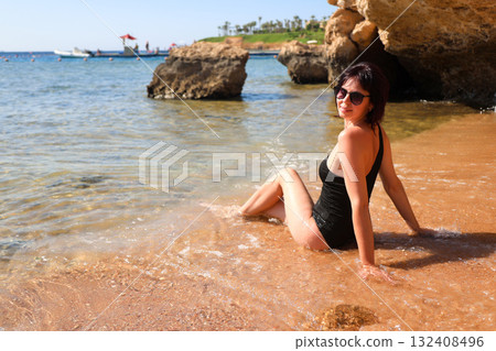 A brunette woman with glasses in a black bathing suit sits on the sand overlooking the sea, view from the back 132408496