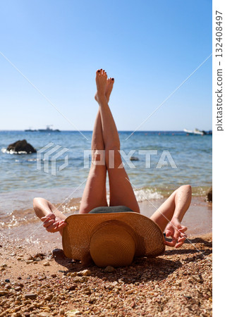 A woman in a wide sun hat lies on the sand overlooking the sea with her legs raised to the sky, enjoying the sun's rays A woman in a wide sun hat lies on the sand overlooking the sea with her legs raised to the sky, enjoying the sun's rays 132408497