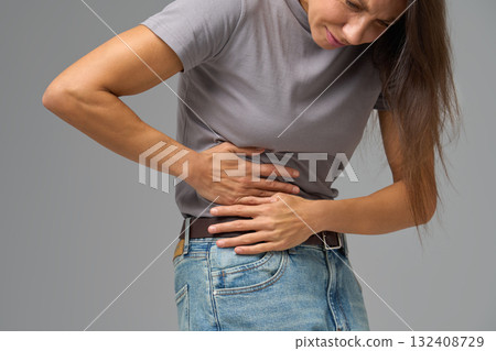 Woman bending sideways and holding abdomen in pain, neutral grey studio setting Woman bending sideways and holding abdomen in pain, neutral grey studio setting 132408729