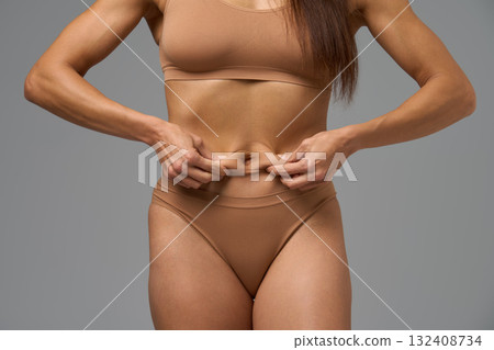 Fit woman in beige underwear holding belly skin showing fitness progress 132408734