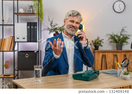 Middle-aged businessman smiles nostalgically speaking on retro cord phone from 90s during work break Middle-aged businessman smiles nostalgically speaking on retro cord phone from 90s during work break 132409148