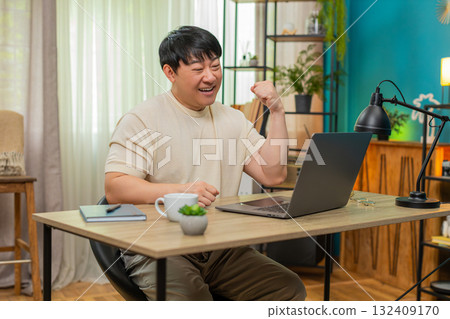 Asian man clenching fist and smiling after completing project on laptop, celebrating career success 132409170