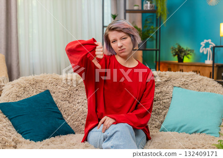 Young woman showing thumbs down gesture negative disapproval disagreement rejection reaction at home Young woman showing thumbs down gesture negative disapproval disagreement rejection reaction at home 132409171