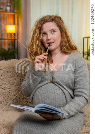 Pregnant young woman writing in diary to do list at home, thoughtful and focused, future planning 132409270