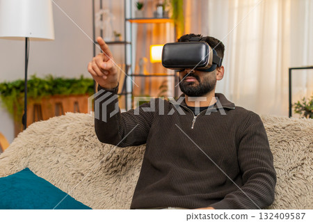 Indian man on sofa wears VR headset at home and reacts with excitement to immersive virtual world Indian man on sofa wears VR headset at home and reacts with excitement to immersive virtual world 132409587