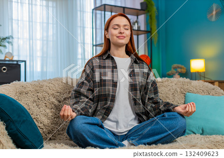 Woman sitting on sofa in lotus pose meditating with closed eyes breathing deeply feeling inner peace Woman sitting on sofa in lotus pose meditating with closed eyes breathing deeply feeling inner peace 132409623
