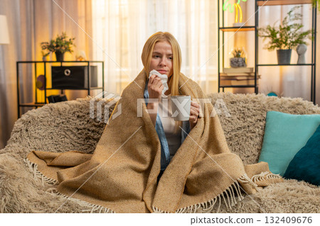 Young woman battling cold at home, wrapped in blanket, drinking hot tea, cozy sofa rest, sniffles Young woman battling cold at home, wrapped in blanket, drinking hot tea, cozy sofa rest, sniffles 132409676