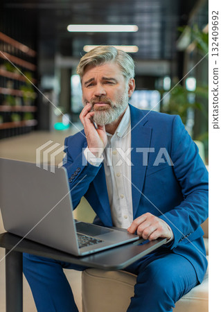 Caucasian businessman in office lobby working on laptop and feeling toothache in modern office lobby Caucasian businessman in office lobby working on laptop and feeling toothache in modern office lobby 132409692