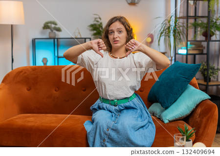 Upset woman showing thumbs down sign gesture, expressing discontent sitting on sofa couch at home 132409694