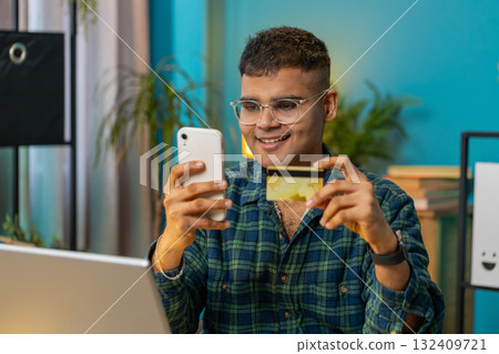 Smiling Indian man using bank credit card and smartphone for online shopping payments at home office 132409721