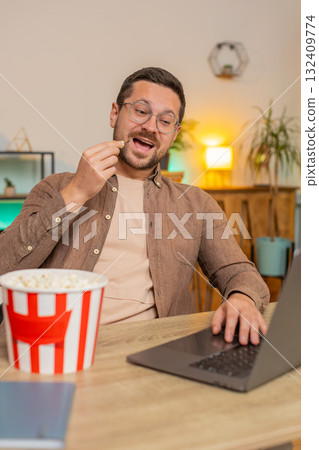Happy young businessman taking break from work eating popcorn and watching movie on laptop at home 132409774