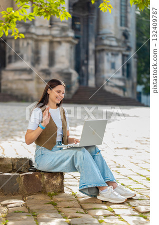 Happy young woman using laptop having video call chat conversation while sitting in city street 132409787
