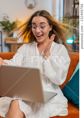 Surprised young woman with windswept hair opening laptop winning lottery sitting on couch at home 132409788