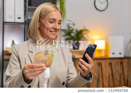 Mature businesswoman counting money euro cash using smartphone calculator app at home office desk 132409789