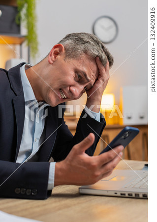Disappointed Caucasian young businessman reading bad news on cellphone sitting at home office table 132409916