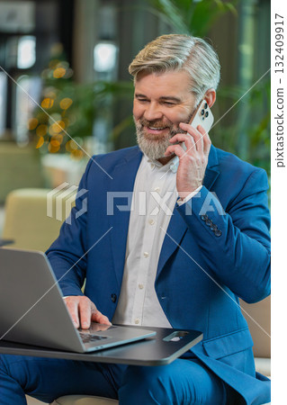 Mature businessman in suit answering to client on mobile phone call, using laptop in office lobby Mature businessman in suit answering to client on mobile phone call, using laptop in office lobby 132409917