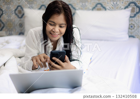 Asian woman using smartphone and laptop on bed for online communication and digital lifestyle at home 132410508