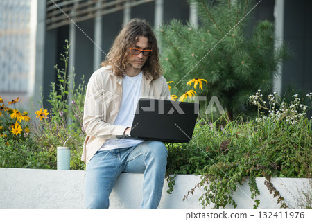 Handsome man, attractive freelancer, brunette sits on bench in urban park, using laptop for remote work. Young businessman or student enjoying work break Outdoors. 132411976