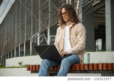 Young man, student or freelancer sits on bench using laptop for remote work at park. Outdoors. Student enjoying work break. 132411977