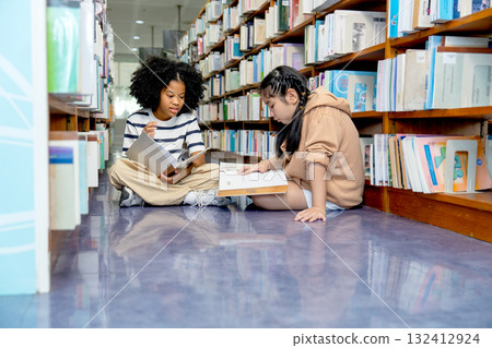 Side view Young two girls sit on floor between bookshelves of public library and enjoy with read and discuss about some books together. 132412924