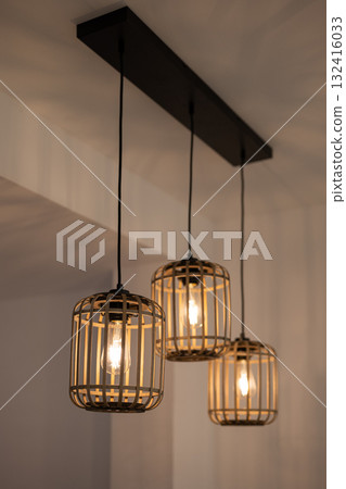 Three pendant lights with a cage design hang from a black ceiling fixture. The lights emit a warm glow, enhancing the modern interior decor. Three pendant lights with a cage design hang from a black ceiling fixture. The lights emit a warm glow, enhancing the modern interior decor. 132416033