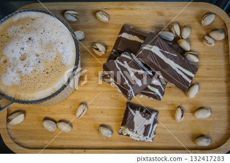 Overhead shot of a coffee cup with foamy coffee accompanied by chocolate pieces topped with nuts. Perfect for food photography, cozy breakfast, desserts, coffee breaks, and lifestyle concepts. 132417283