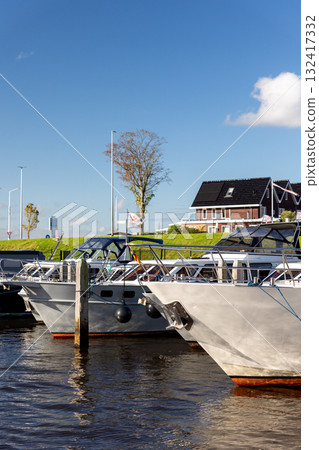 Modern luxury boats moored at Zwartsluis Netherlands canal marina bright sunlight modern houses trees background. Calm water reflects the vessels and blue sky, creating a serene Dutch coastal scene 132417332