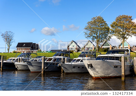 Modern luxury boats moored at Zwartsluis Netherlands canal marina bright sunlight modern houses trees background. Calm water reflects the vessels and blue sky, creating a serene Dutch coastal scene Modern luxury boats moored at Zwartsluis Netherlands canal marina bright sunlight modern houses trees background. Calm water reflects the vessels and blue sky, creating a serene Dutch coastal scene 132417334