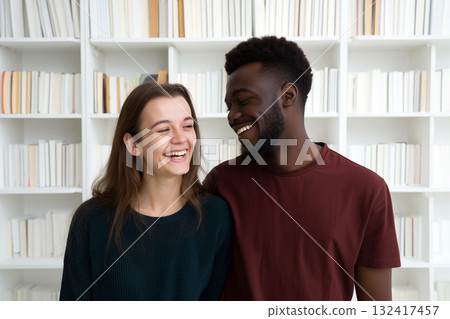 Happy couple laughing together in front of a white bookshelf, enjoying a lighthearted moment indoors Happy couple laughing together in front of a white bookshelf, enjoying a lighthearted moment indoors 132417457