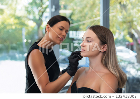 Makeup artist applies makeup to client in a bright studio with natural light and greenery outside Makeup artist applies makeup to client in a bright studio with natural light and greenery outside 132418302