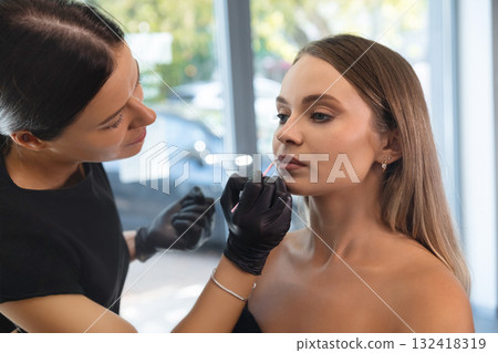 Makeup artist applies lip color to model during a beauty session in a bright studio in the morning Makeup artist applies lip color to model during a beauty session in a bright studio in the morning 132418319