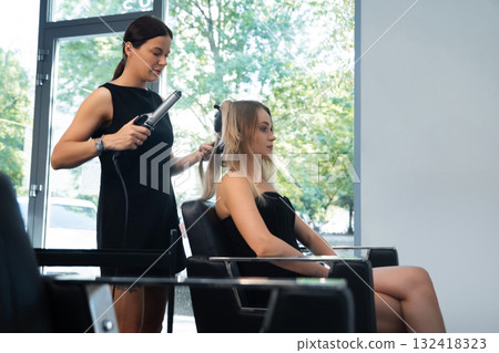 A hair stylist styles a client hair in a contemporary salon with large windows letting in natural light. The atmosphere feels relaxing and professional A hair stylist styles a client hair in a contemporary salon with large windows letting in natural light. The atmosphere feels relaxing and professional 132418323