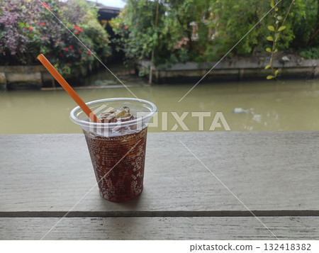 Iced drink plastic cup orange straw ice cubes wooden table river outdoor refreshing mood 132418382