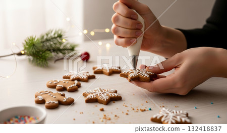 Close-up of hands decorating homemade gingerbread cookies with white icing in a cozy Christmas setting with lights. 132418837