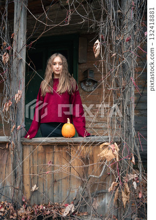 Young woman in a witch hat and red sweater stands near a rustic wooden house with a pumpkin on the railing, surrounded by dry vines Young woman in a witch hat and red sweater stands near a rustic wooden house with a pumpkin on the railing, surrounded by dry vines 132418881