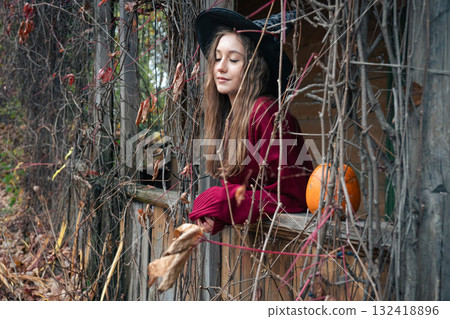 Young woman in a witch hat and burgundy sweater leans out of a rustic wooden window surrounded by dry vines and a pumpkin, Halloween atmosphere Young woman in a witch hat and burgundy sweater leans out of a rustic wooden window surrounded by dry vines and a pumpkin, Halloween atmosphere 132418896