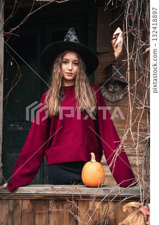 Young woman in a witch hat and burgundy sweater poses on a rustic wooden porch with a pumpkin, surrounded by dry autumn vines Young woman in a witch hat and burgundy sweater poses on a rustic wooden porch with a pumpkin, surrounded by dry autumn vines 132418897