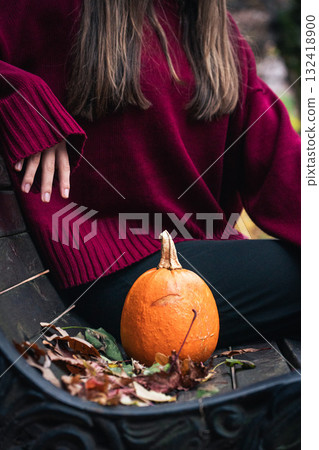 Close-up of a small orange pumpkin on a park bench next to a person in a burgundy sweater, surrounded by autumn leaves Close-up of a small orange pumpkin on a park bench next to a person in a burgundy sweater, surrounded by autumn leaves 132418900