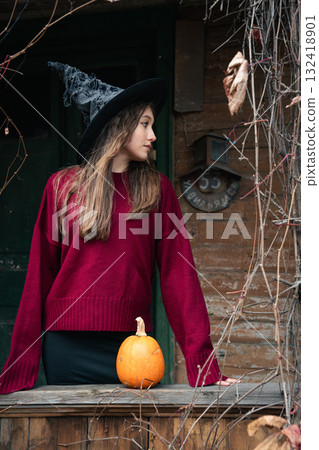 Young woman in a witch hat and red sweater stands near a rustic wooden house with a pumpkin on the railing, surrounded by dry vines 132418901