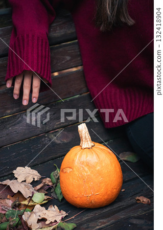 Close-up of a small orange pumpkin on a park bench next to a person in a burgundy sweater, surrounded by autumn leaves Close-up of a small orange pumpkin on a park bench next to a person in a burgundy sweater, surrounded by autumn leaves 132418904