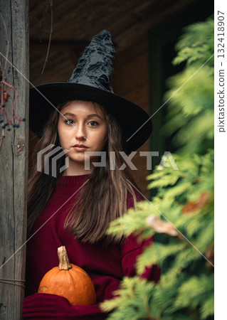 Young woman in a burgundy sweater stands on the wooden porch of an old house holding a small pumpkin, surrounded by autumn leaves and greenery Young woman in a burgundy sweater stands on the wooden porch of an old house holding a small pumpkin, surrounded by autumn leaves and greenery 132418907