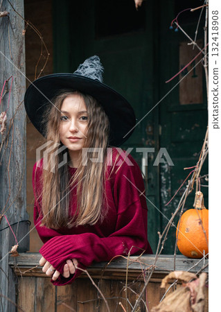 Young woman in a witch hat and burgundy sweater leans out of a rustic wooden window surrounded by dry vines and a pumpkin, Halloween atmosphere 132418908